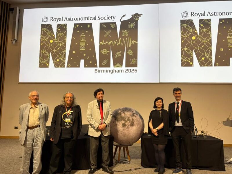 Four men and 1 woman on a stage with a big globe of a moon between them and a banner overhead.