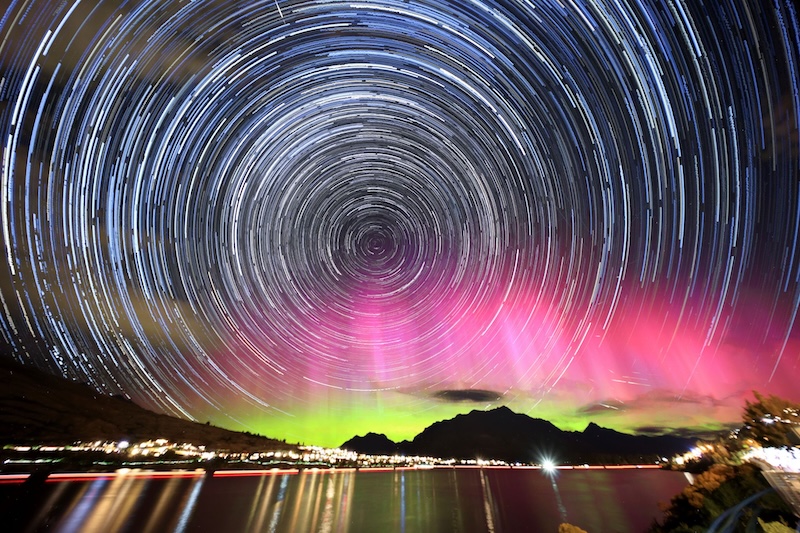 Star trails with aurora lighting up the horizon above a distant city overlooking a lake.