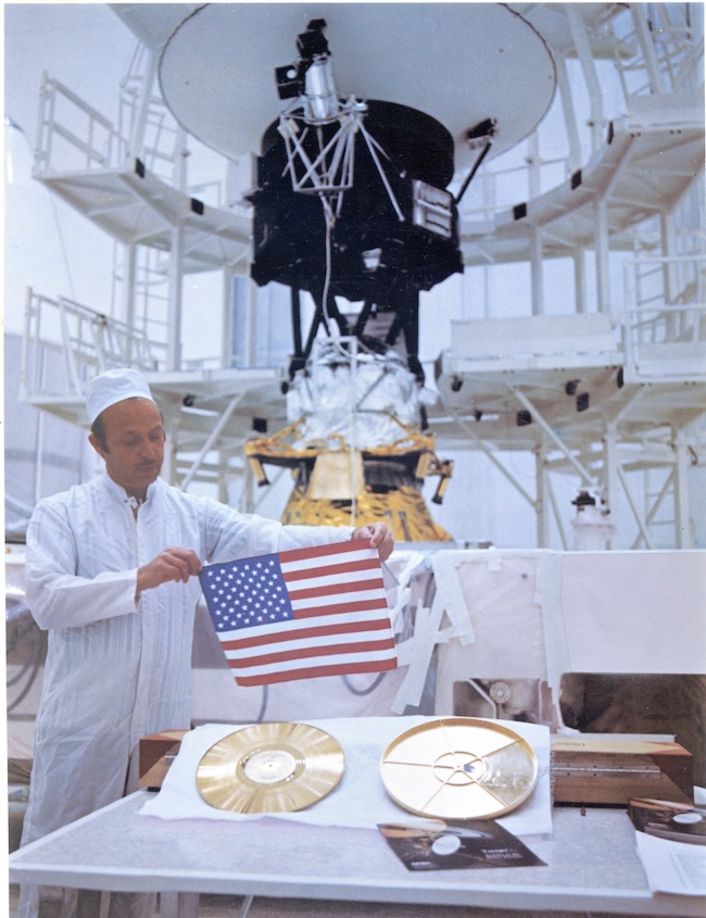 Voyager 2: Man in white holding a small American flag above 2 golden circular disks, spacecraft in background.
