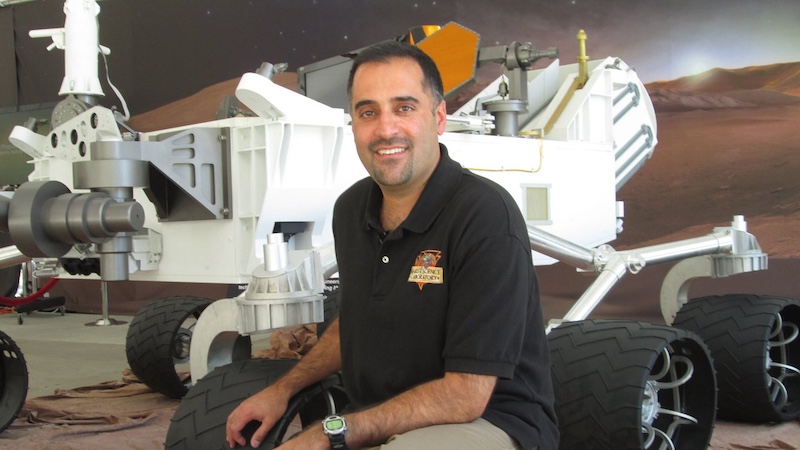 Man with short hair and shirt with mission logo sitting in front of a full-scale model of a Mars rover.