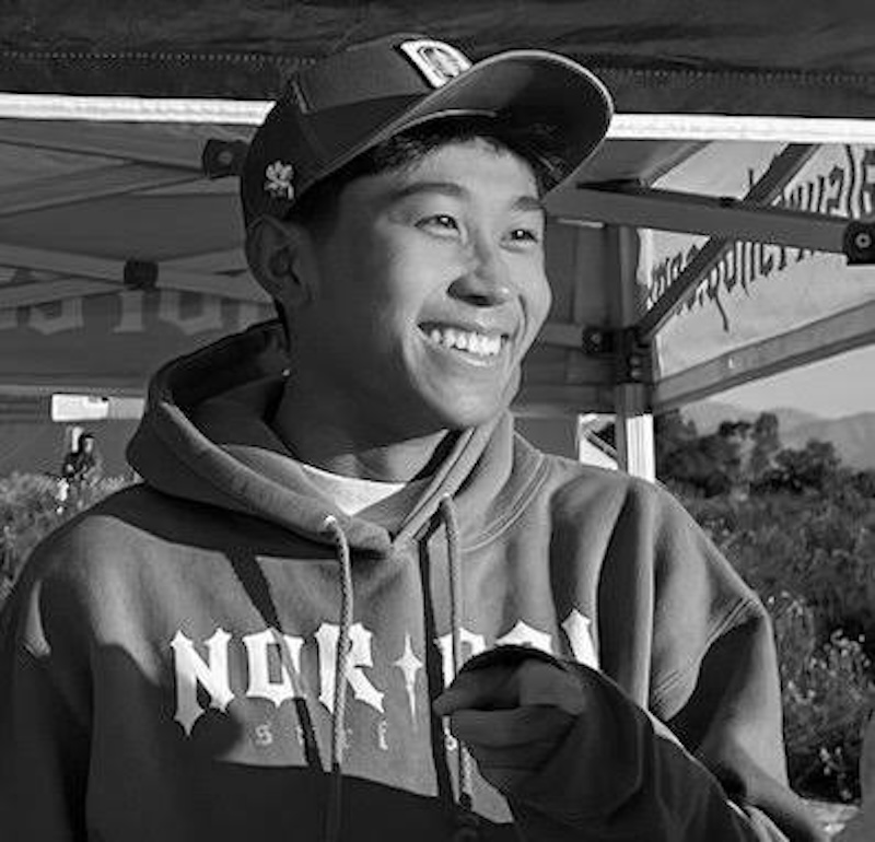 Black and white photo of smiling Asian man wearing a ball cap and hoodie.