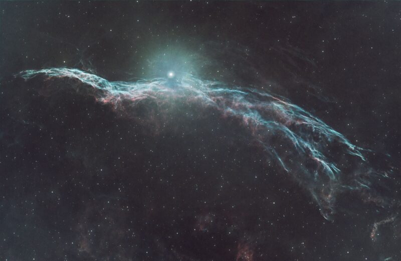 A large arc of nebulosity in pink and baby blue, with numerous stars immersed within and in the surroundings.