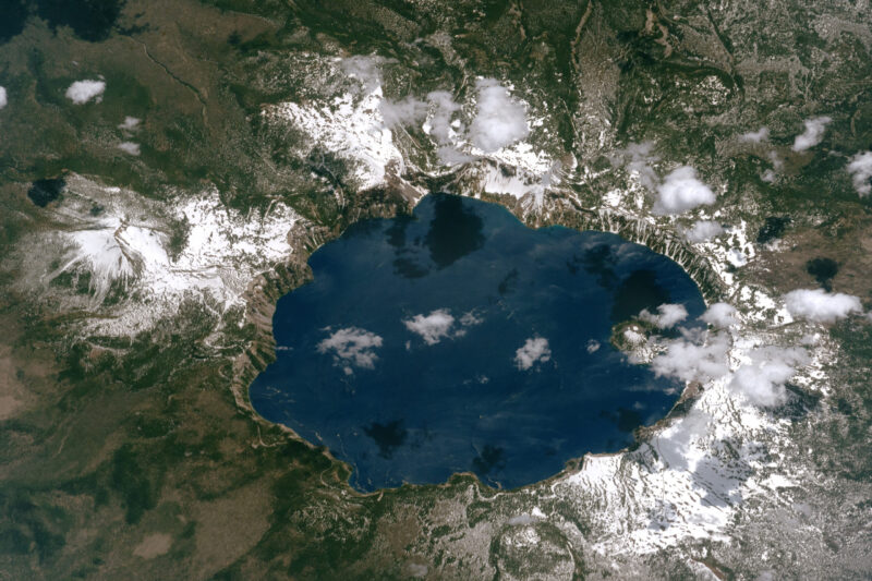 A large blue lake with a volcanic-looking mountain inside and some white scattered around the edges of the water-filled crater.