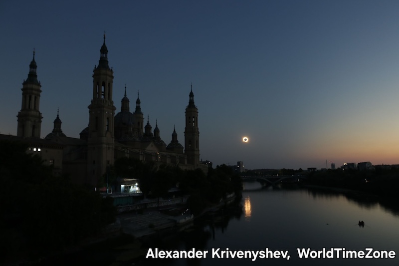 Distant eclipsed sun, also reflected in a river, with Gothic church spires in the foreground.