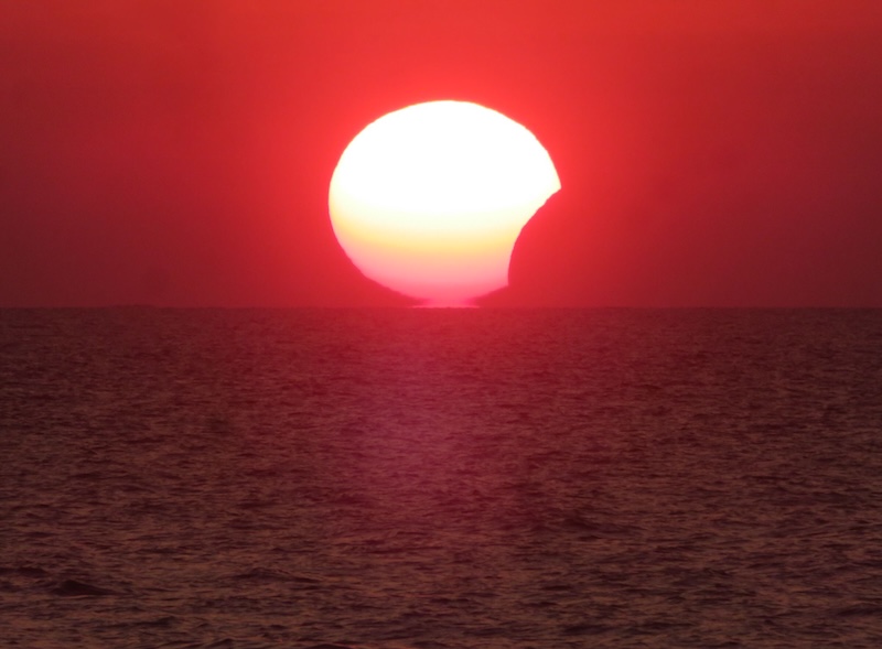 Bright solar crescent over a reddish sky, descending over the ocean.