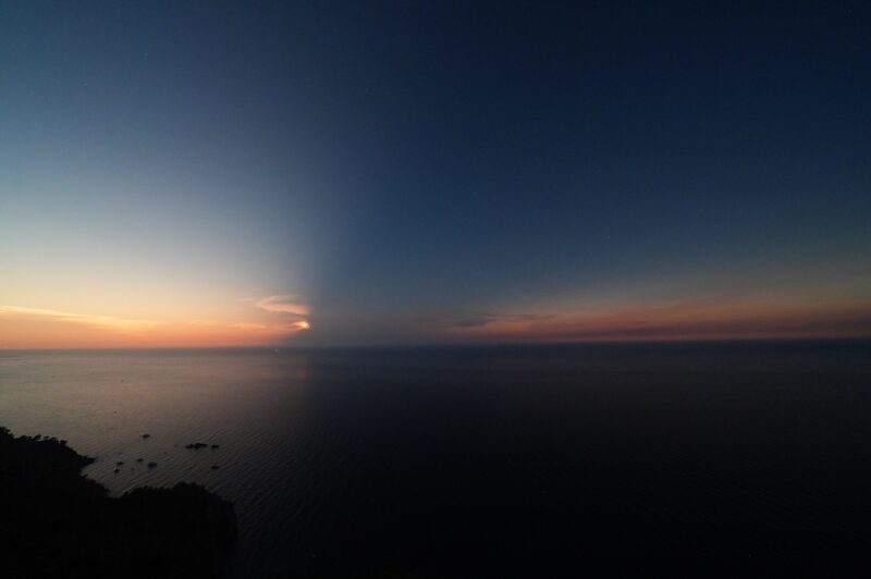 A coastal view towards the Mediterranean, with deep shadows over water and sky, and the orange tint of sunset.