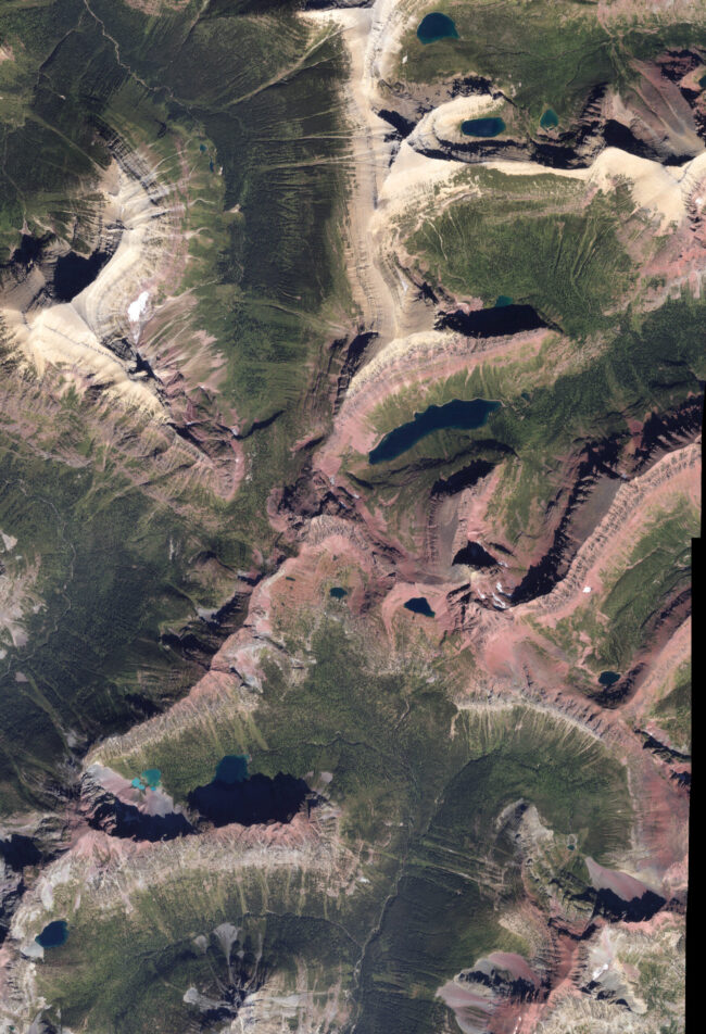 View of green landscape with reddish rocks and many steep slopes and some bright blue lakes.