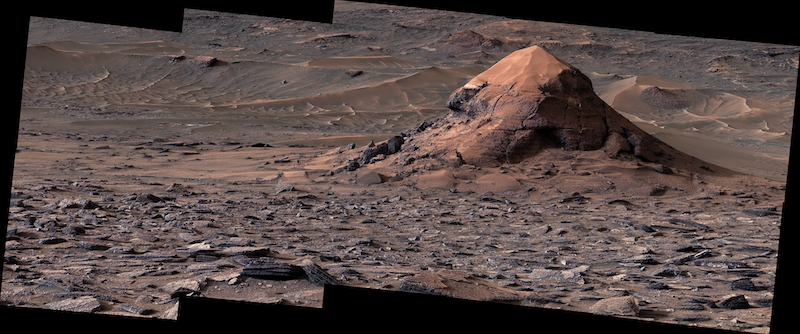 Conical rocky mound with sand on the peak, in brownish, choppy terrain.