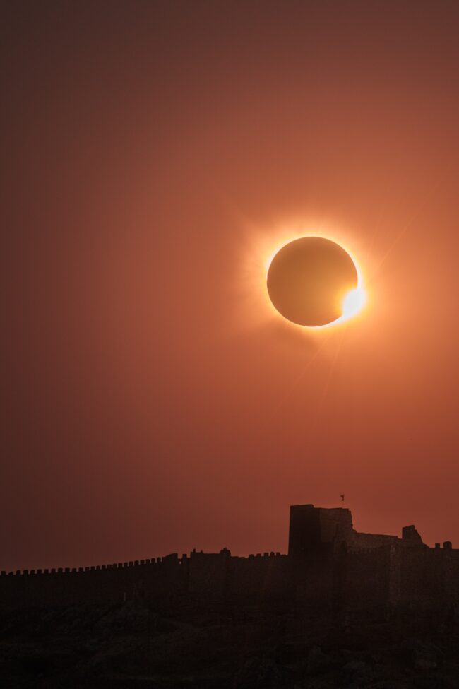 A black disk, the sun, surrounded by a luminous aura and a bright of golden light, over an orange-colored sky.