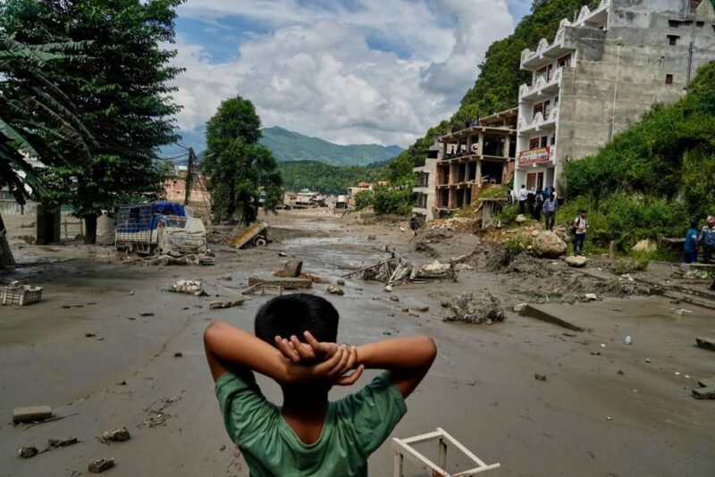 A boy stands with his arms behind his head, facing a street lined with damaged multistory buildings.