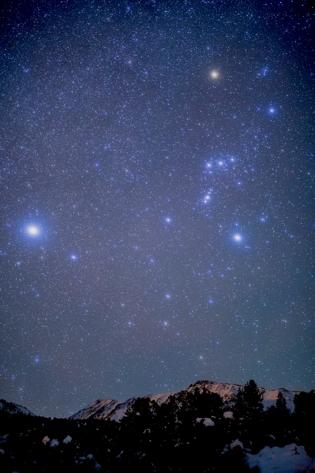 Distant mountains under a starry sky, including high Orion and a very bright dot to the left and a bit lower than Orion.