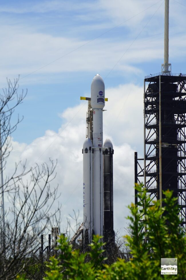 A tall rocket with cylindrical boosters attached, standing against fluffy clouds in a blue sky.