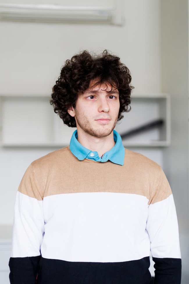 Young man with dark, curly hair, wearing a striped sweater with a blue shirt collar sticking out.
