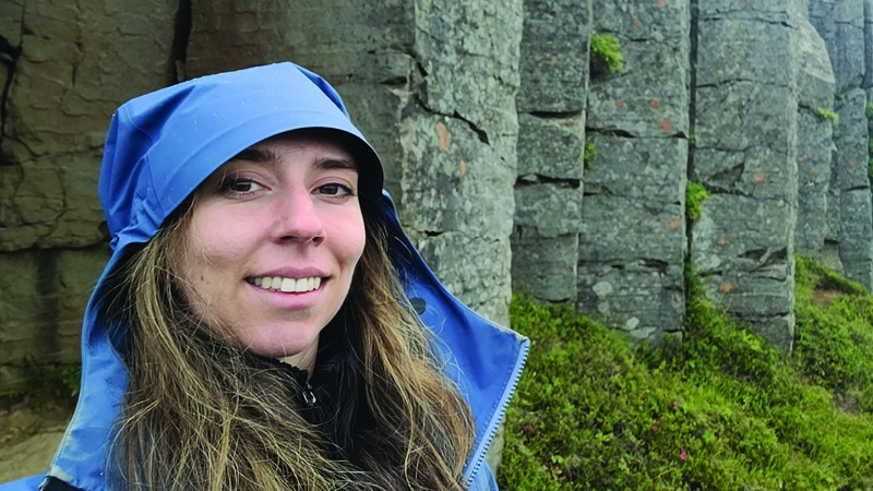 Smiling woman wearing a blue rain cape and hoodie, with stone columns behind her.