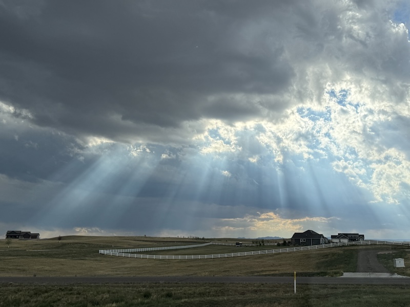 Streamers of crepuscular rays coming out of dark clouds over a few houses and prairie land.