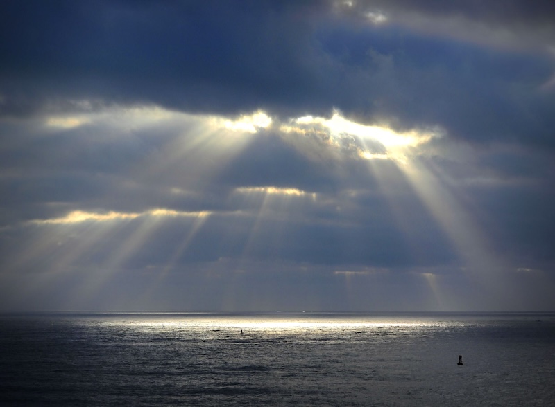 Streamers of crepuscular rays coming out of heavy clouds over the ocean.