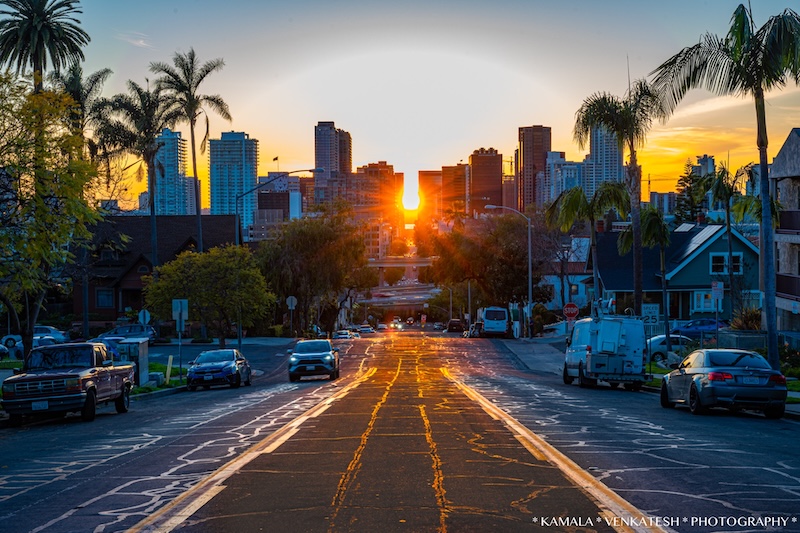 Setting sun peaking through distant high-rise buildings behind a four-lane city street with cars.