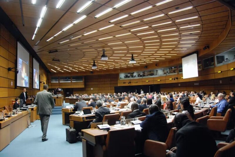 A semicircular room with dozens of people sitting at desks watching a big screen up front.