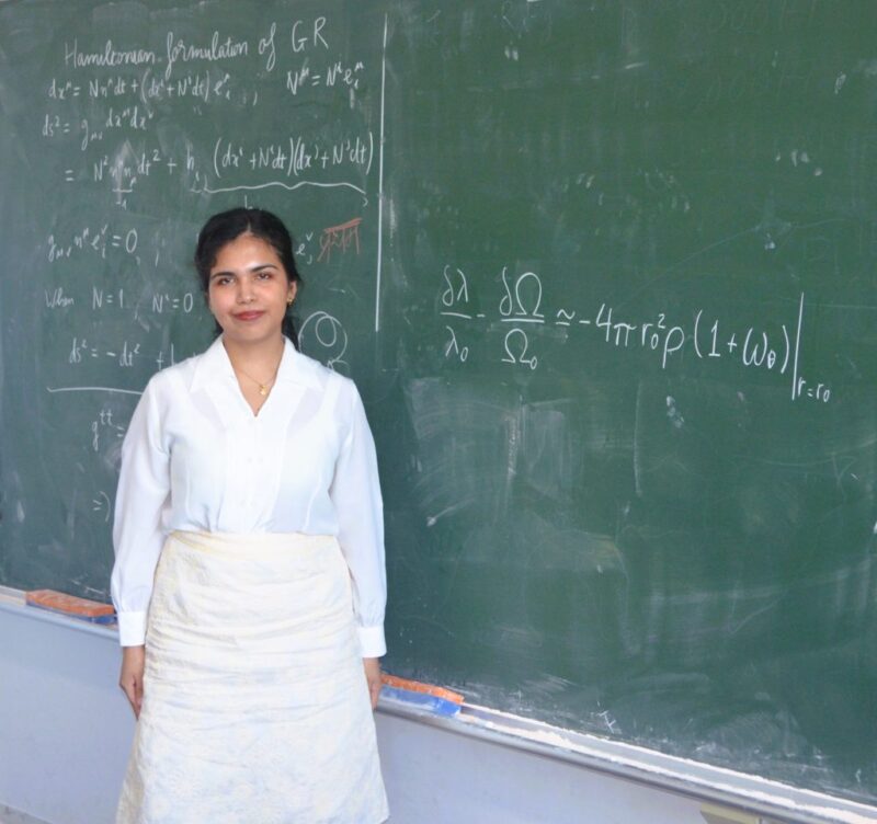A black-haired woman wearing white stands beside a chalkboard with math equations on it.