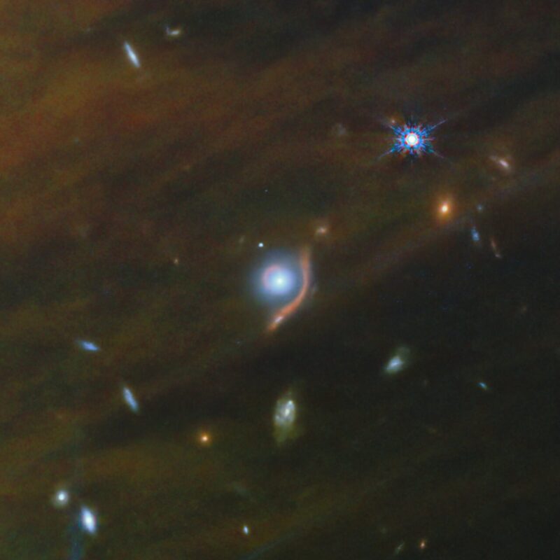 Small, distant galaxies with a larger bright one in the middle with a reddish curved line partway around it.