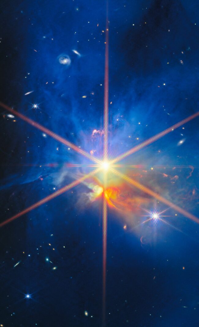 BBright star at center, wispy blue and orange dust clouds, and many small, distant galaxies.