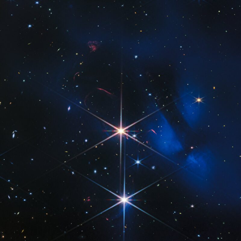 Starfield with 2 very bright stars ad distant galaxies, plus faint blue clouds and thin red streaks.