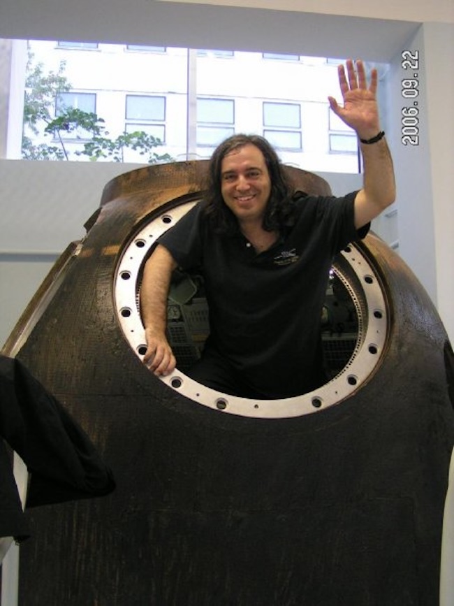 Man standing in porthole opening in a dark object, smiling and waving his hand.