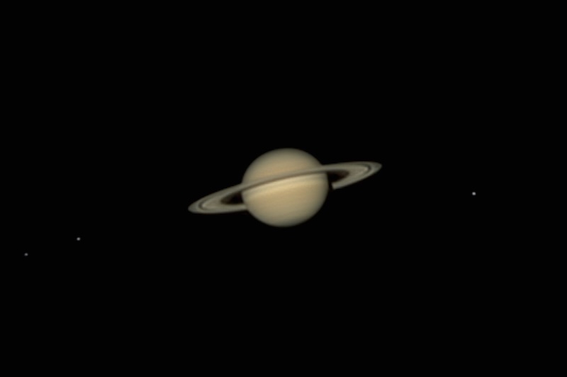 Saturn through a telescope with the rings slightly tilted upward. Nearby are three dots that are the moons Dione, Tethys and Rhea.