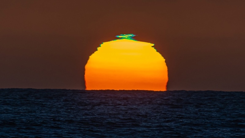 The sun, partly set, with a green glow at its top.
