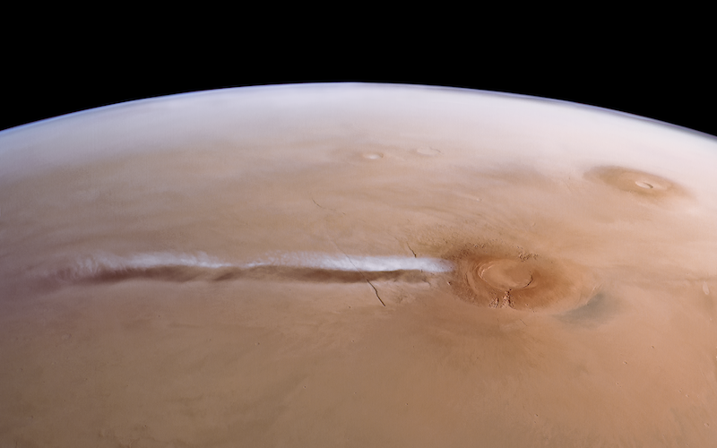 Long, white cloud extending from a volcano on a brownish planet, seen from above.