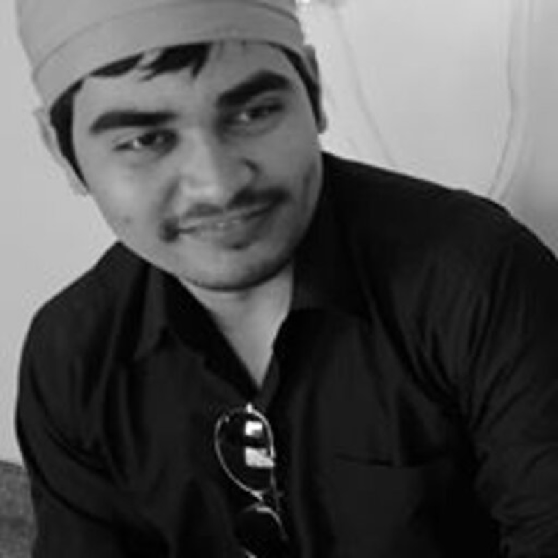 Black and white photo of a smiling man wearing a cap and dark jacket. His sunglasses are hanging from his shirt neckline.