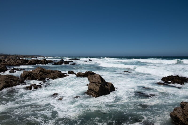 A blue sky over a shore scattered with big rocks with strong waves crashing against them.