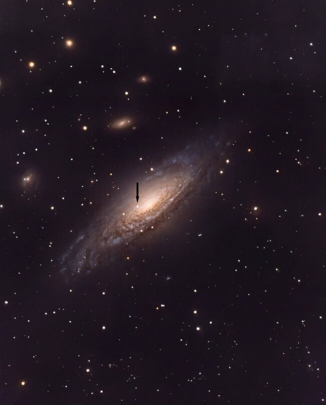 Large, yewllowish galaxy, a spiral seen almost edge-on, with a black arrow and numerous foreground stars.
