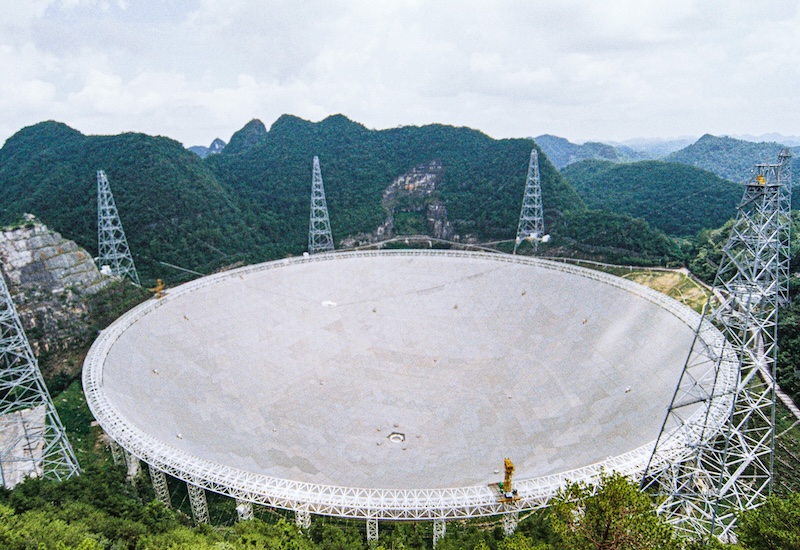 Huge round radio dish, with tall radio towers around the edges, near mountains.