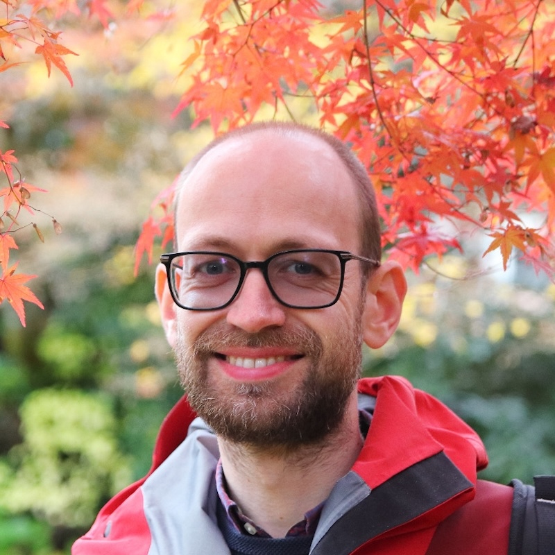 Smiling, partly bald man with beard and black eyeglasses, seen in front of autumn leaves.