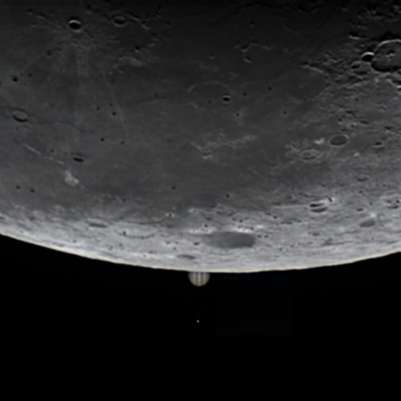 Limb of the cratered moon above, intersecting with a tiny, distant Jupiter, and a dot below it for its largest moon.