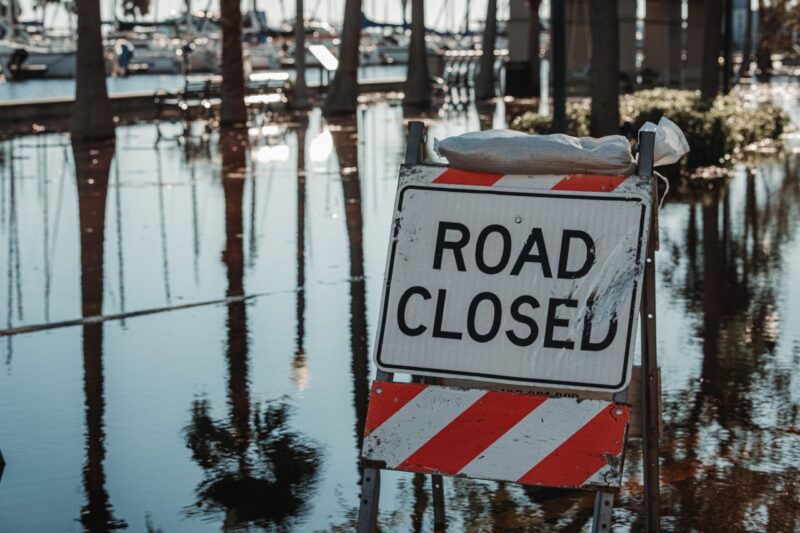 Road, filled with standing water reflecting trees, and in the foreground a sign with road closed on it.