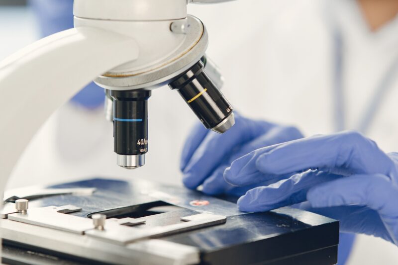Close-up view of a white laboratory microscope operated by a person wearing blue nitrile gloves.
